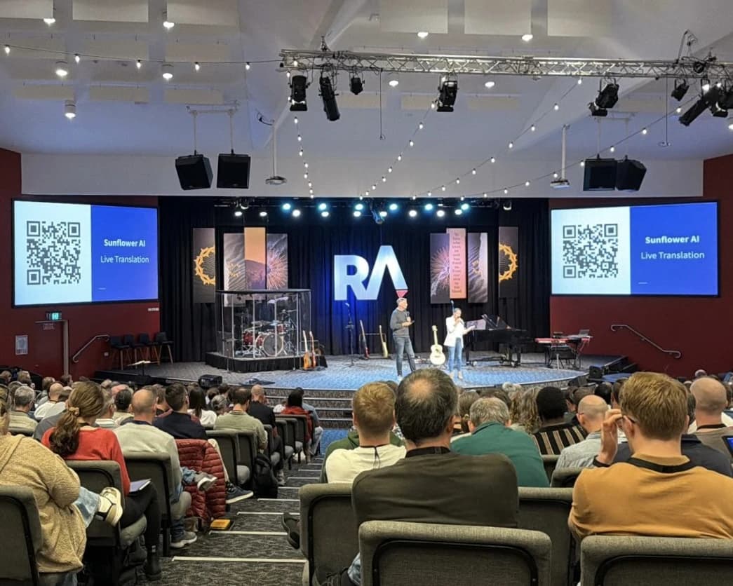 A packed auditorium at Reach Australia's National Conference, with screens either side of the stage showing Sunflower AI live translation QR codes